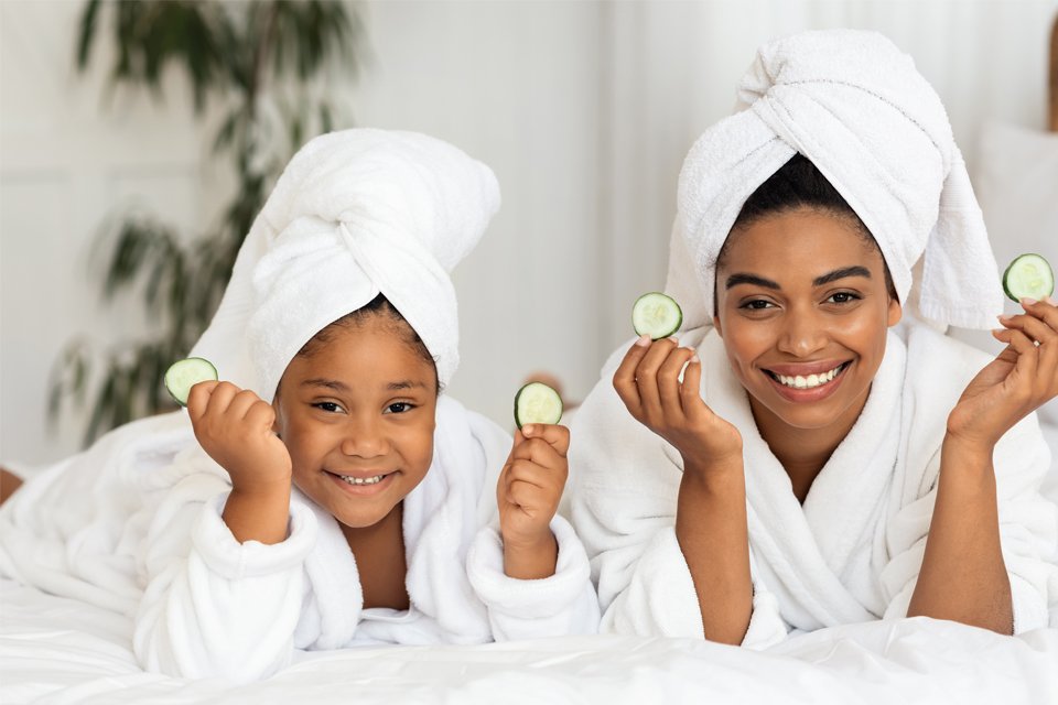 A mother and daughter in robes hold cucumber slices near their faces on a spa day.